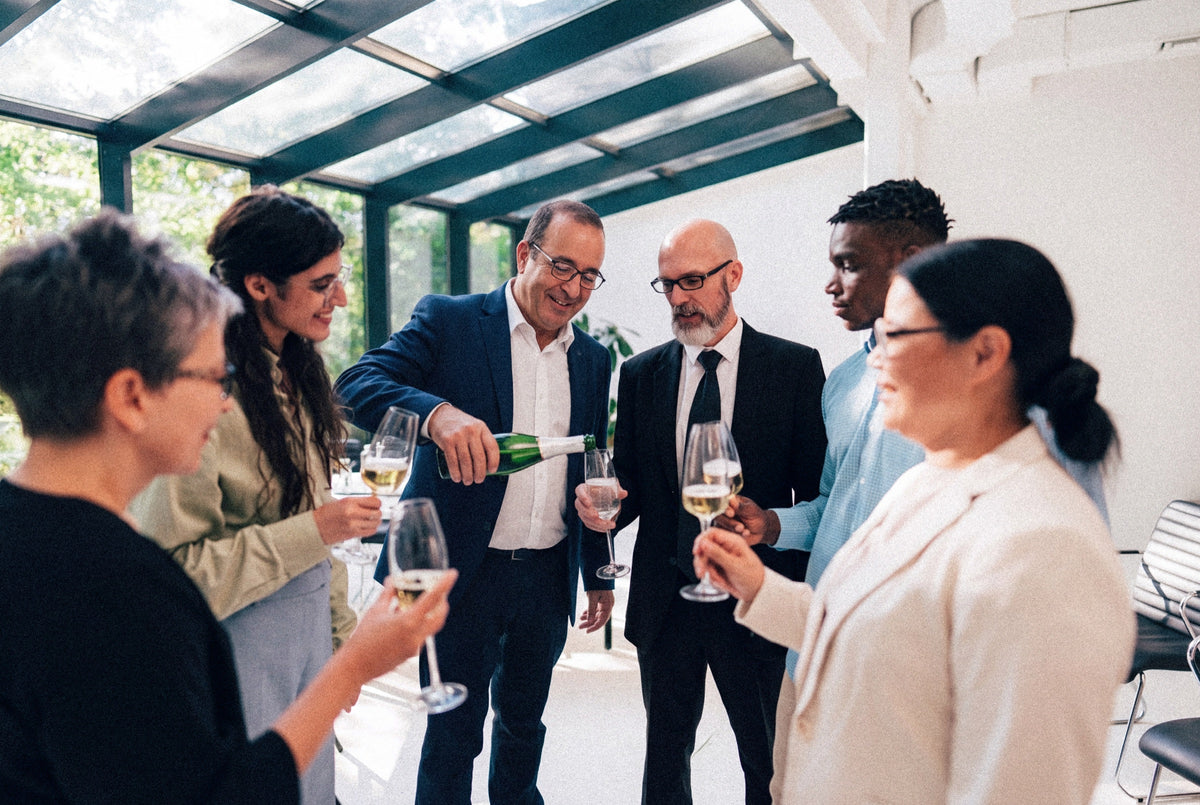 Peter pours sparkling wine for a group of guests in a sunlit glass-roofed room